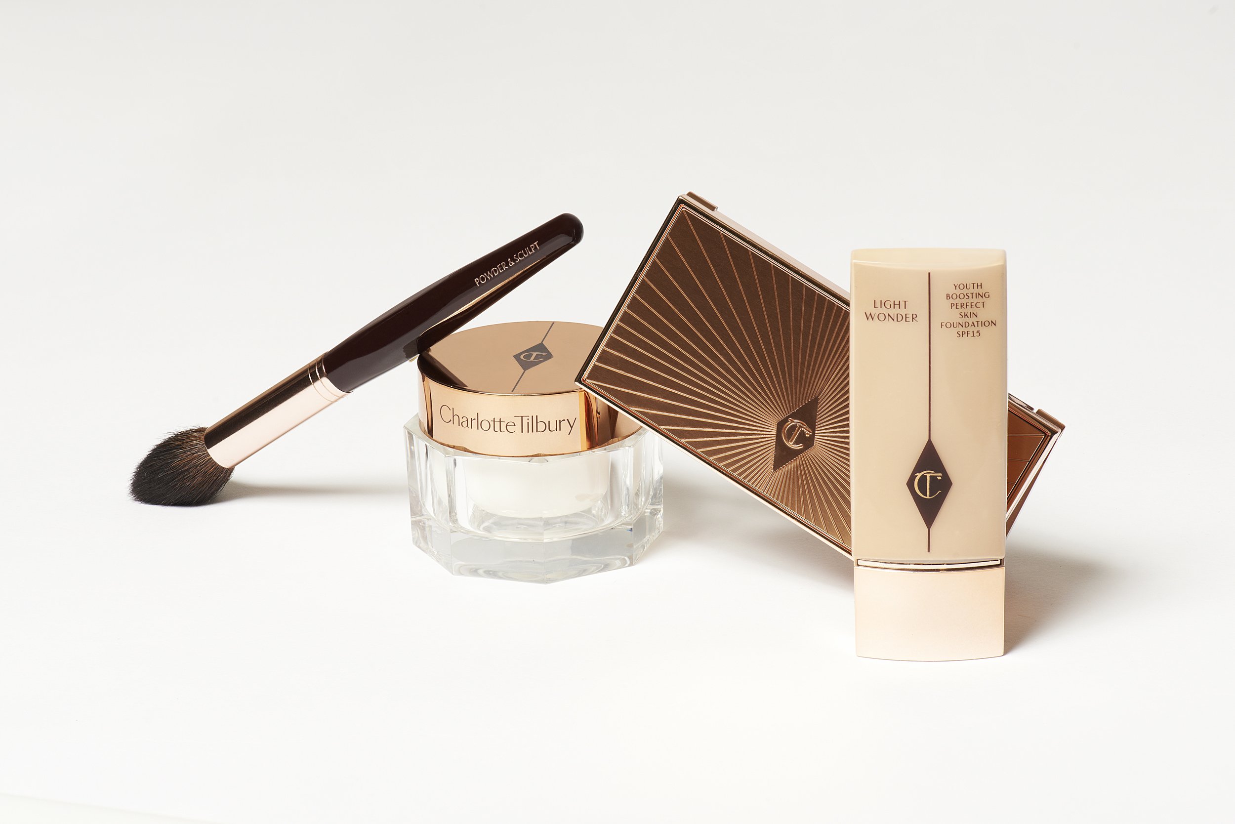 Charlotte Tilbury makeup products including powder, foundation, and a brush on a white background. Beauty and cosmetics still life.