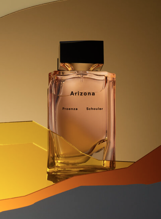 Still life of a translucent glass Proenza Schouler Arizona perfume bottle with a black cap on a gold geometric surface.