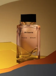 A rectangular glass perfume bottle with a dark cap resting on a geometric reflective yellow and orange surface against a warm gold backdrop.