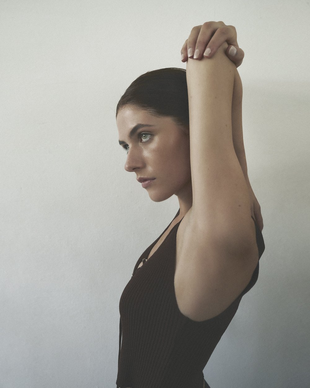Portrait of a young adult woman in a dark brown ribbed tank top, indoors in front of a white wall with soft, natural lighting.