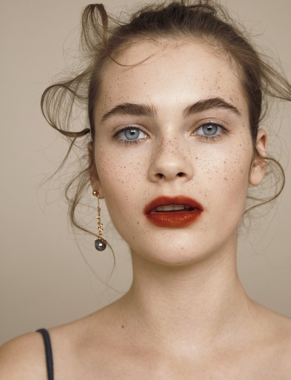 Close-up portrait of a woman with freckles, red lipstick, and an earring. She has light eyes, and her brown hair is styled up.