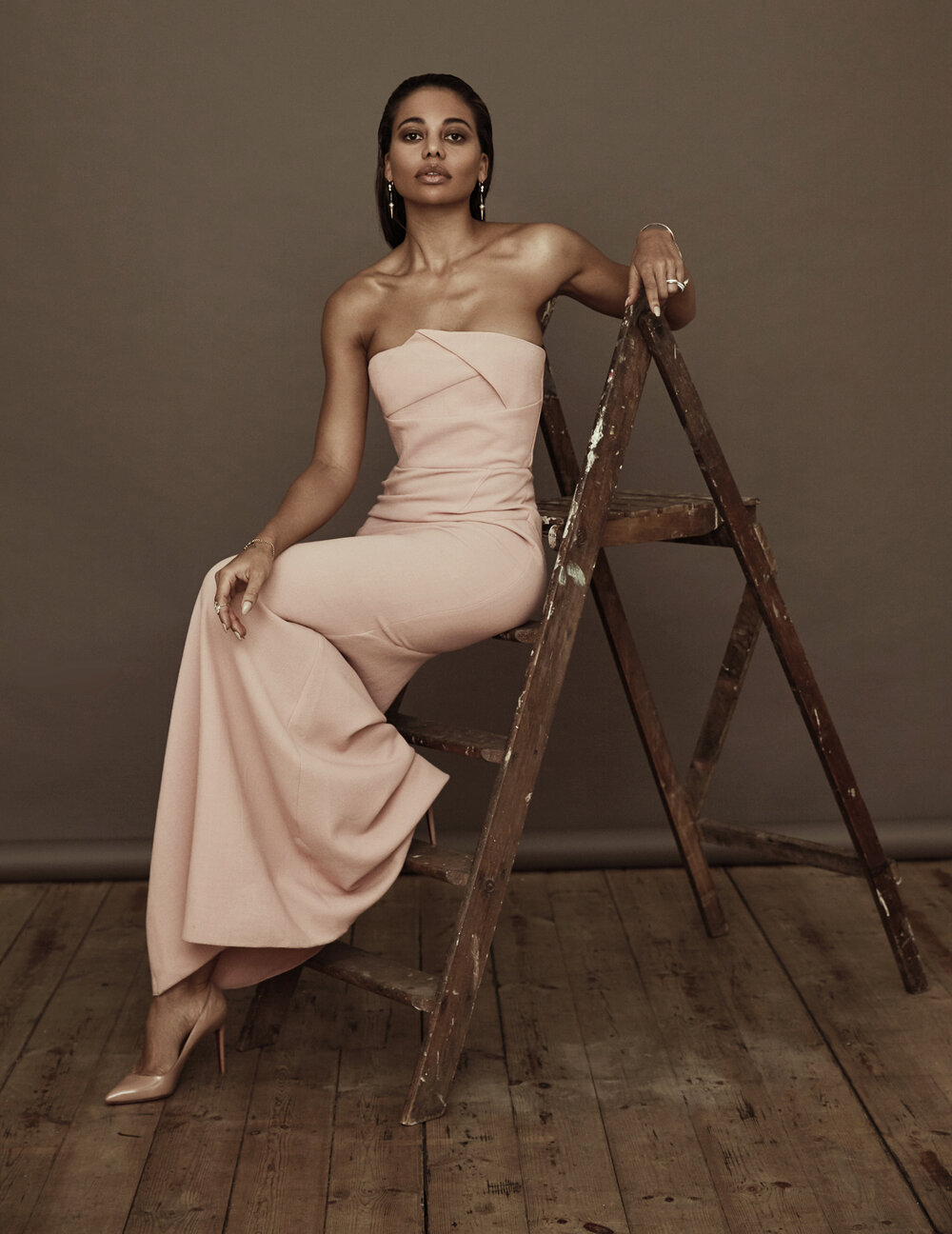 A young woman in a peach-colored strapless jumpsuit poses gracefully on a wooden ladder indoors against a neutral grey background.
