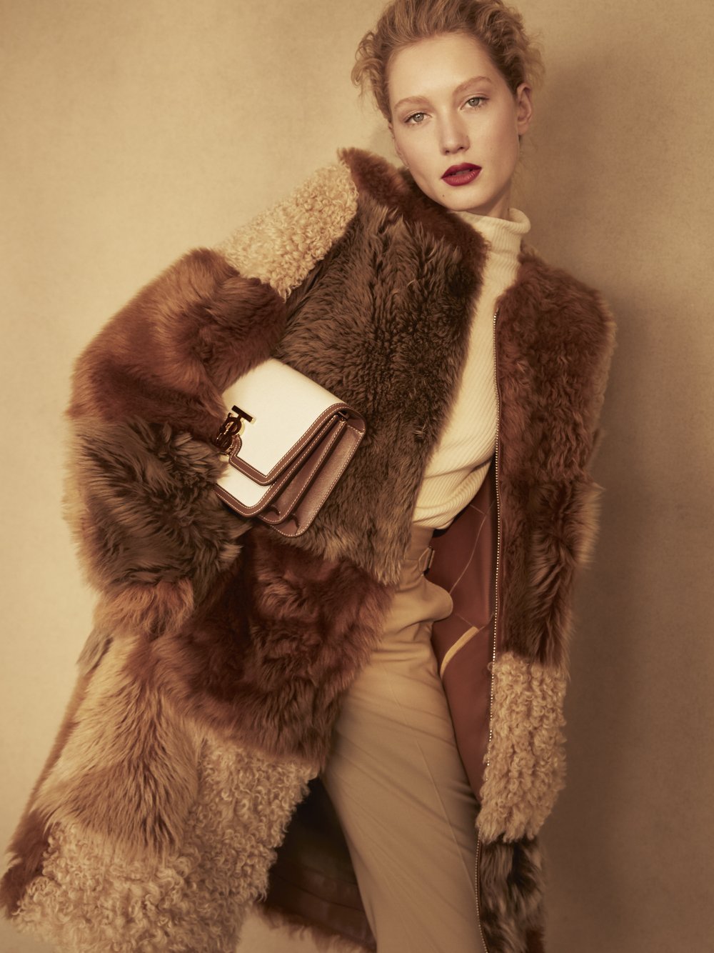 Fashionable adult woman poses in a studio wearing a luxurious brown fur coat, cream turtleneck, and holding a stylish white handbag.