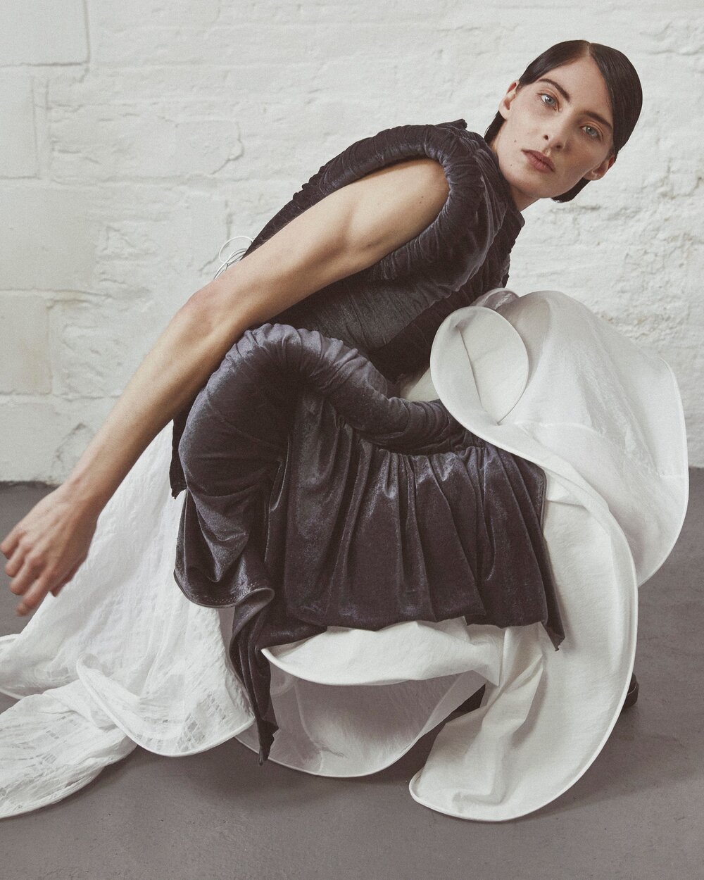 An adult female model poses in a crouched position wearing a dark-colored fashion dress with a large, flowing white fabric accent.