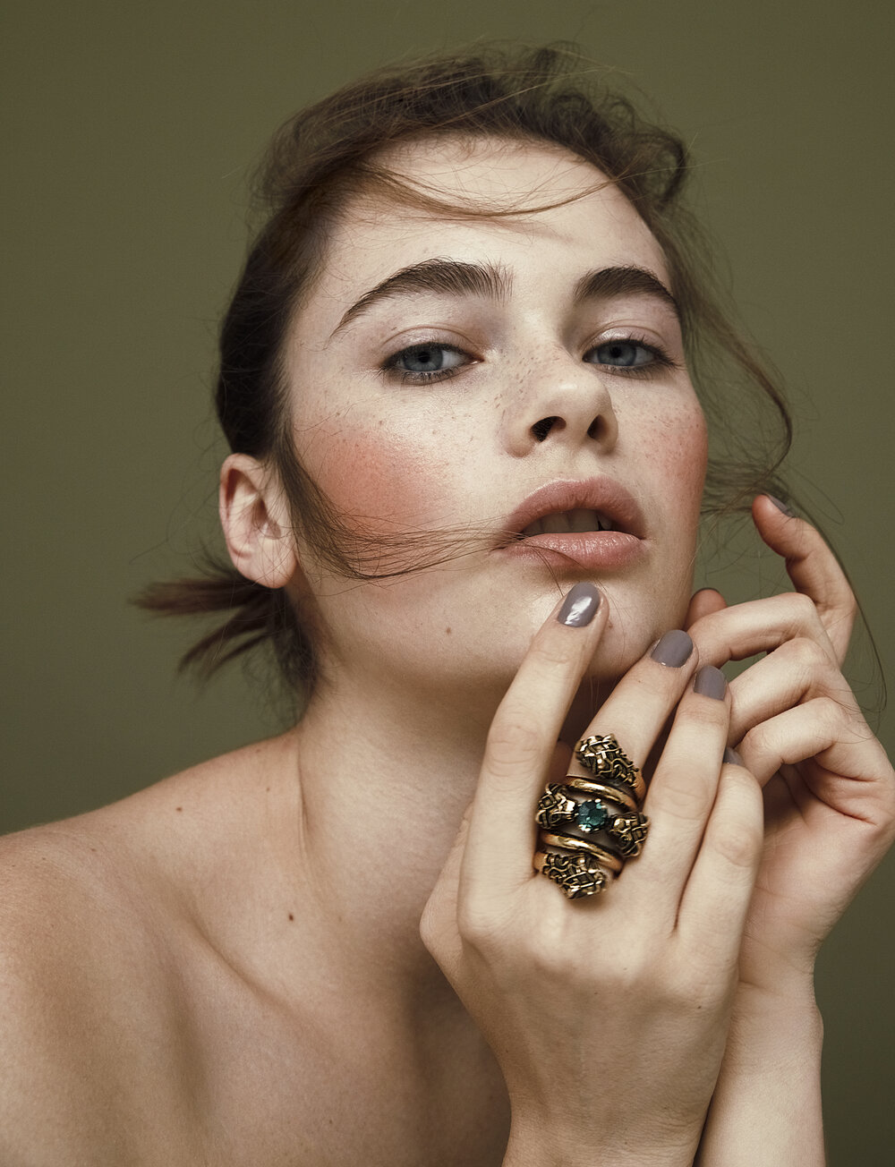Close-up studio portrait of a woman with light skin wearing a large gold statement ring against a muted green backdrop.