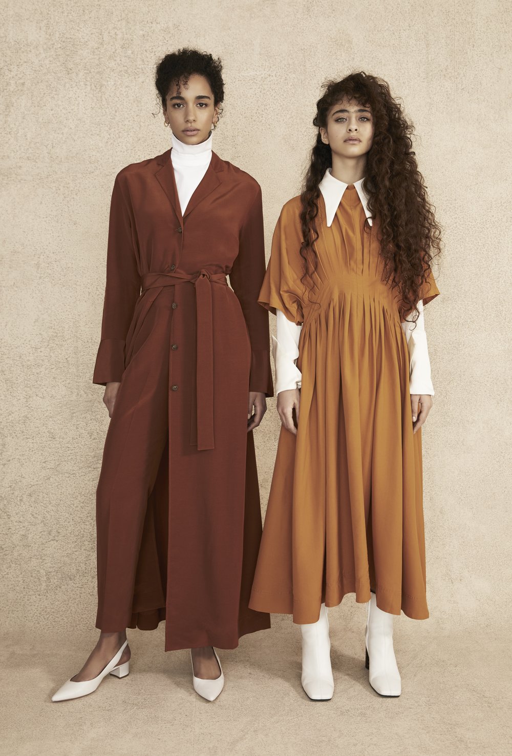 Two women model earth-tone dresses and white shoes in a full shot against a mottled beige backdrop, creating a modern fashion editorial.
