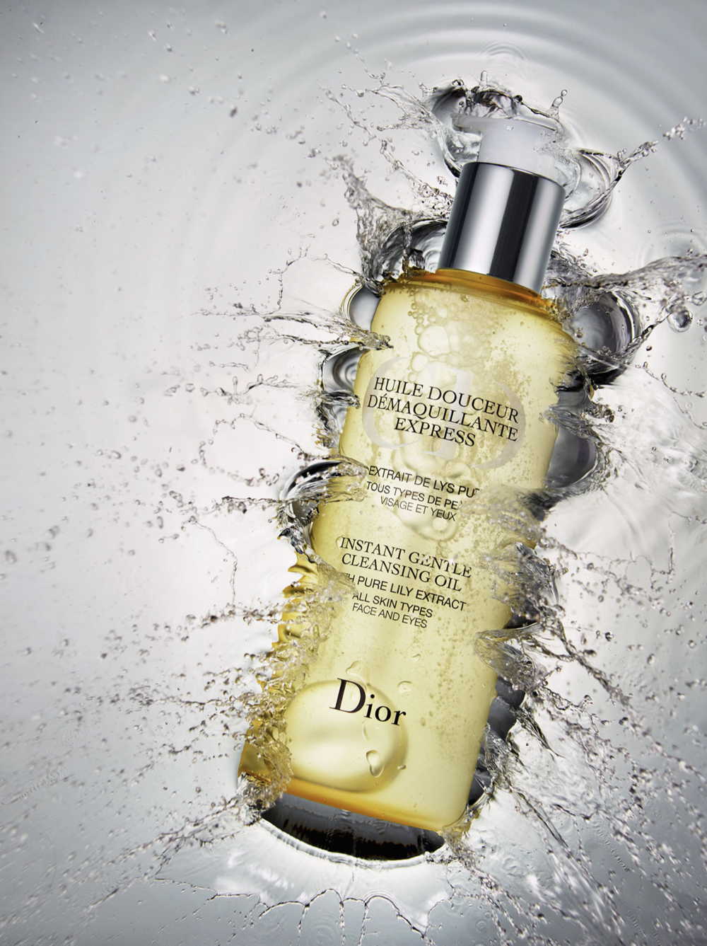 Dior cleansing oil bottle, clear and yellow liquid, surrounded by splashing water on a gray background in a beauty product shot.
