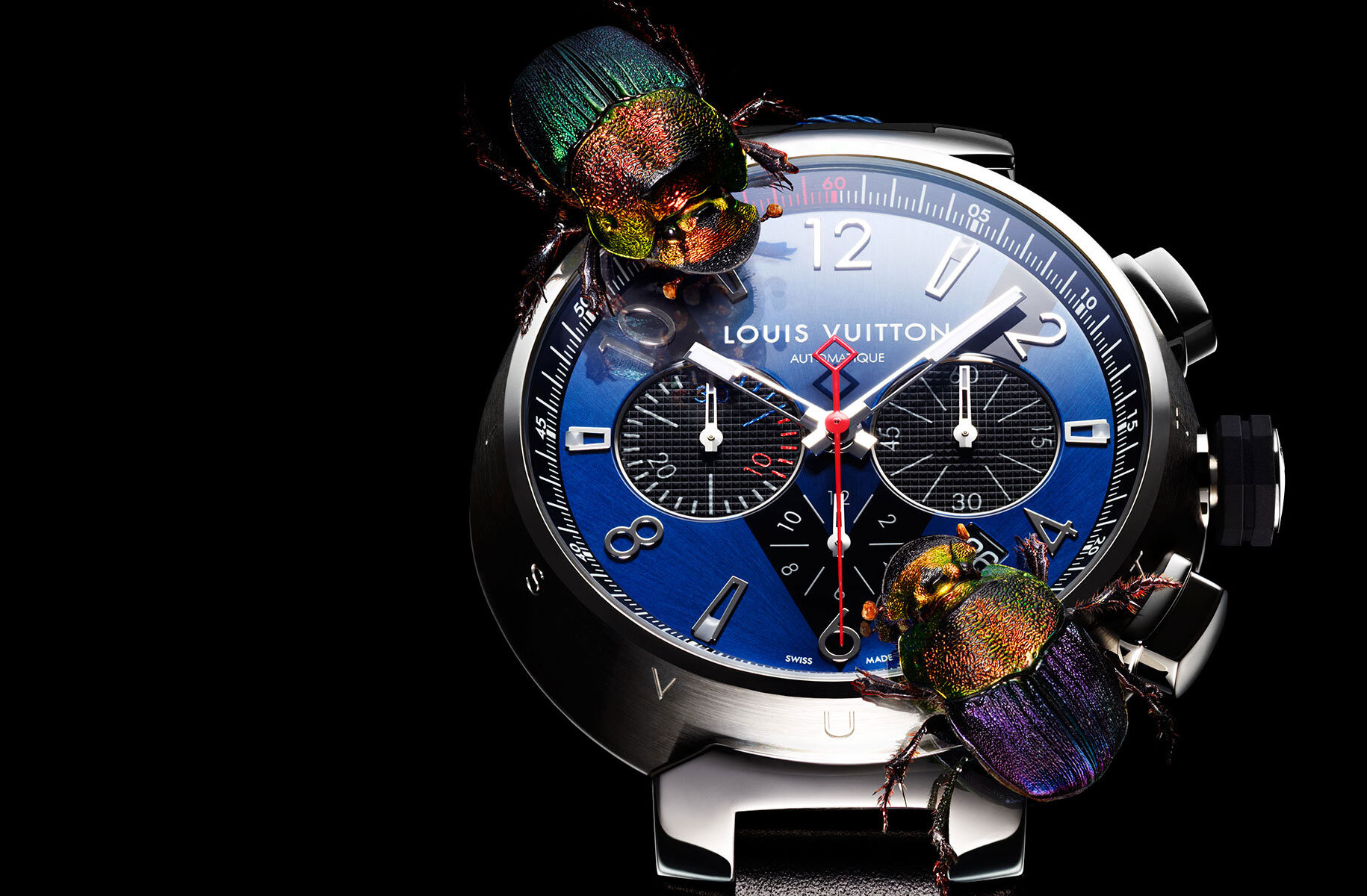 Close-up of a Louis Vuitton watch with a blue face, silver casing, and two iridescent beetles on its surface, set against a dark background.