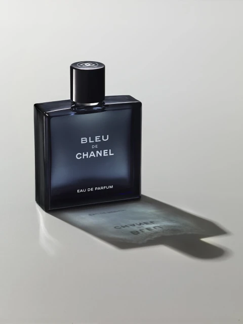 Close-up studio shot of a blue glass Chanel perfume bottle on a white surface, casting a shadow with reflected text.