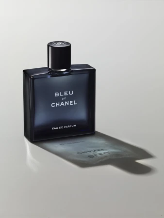 Close-up studio shot of a blue glass Chanel perfume bottle on a white surface, casting a shadow with reflected text.