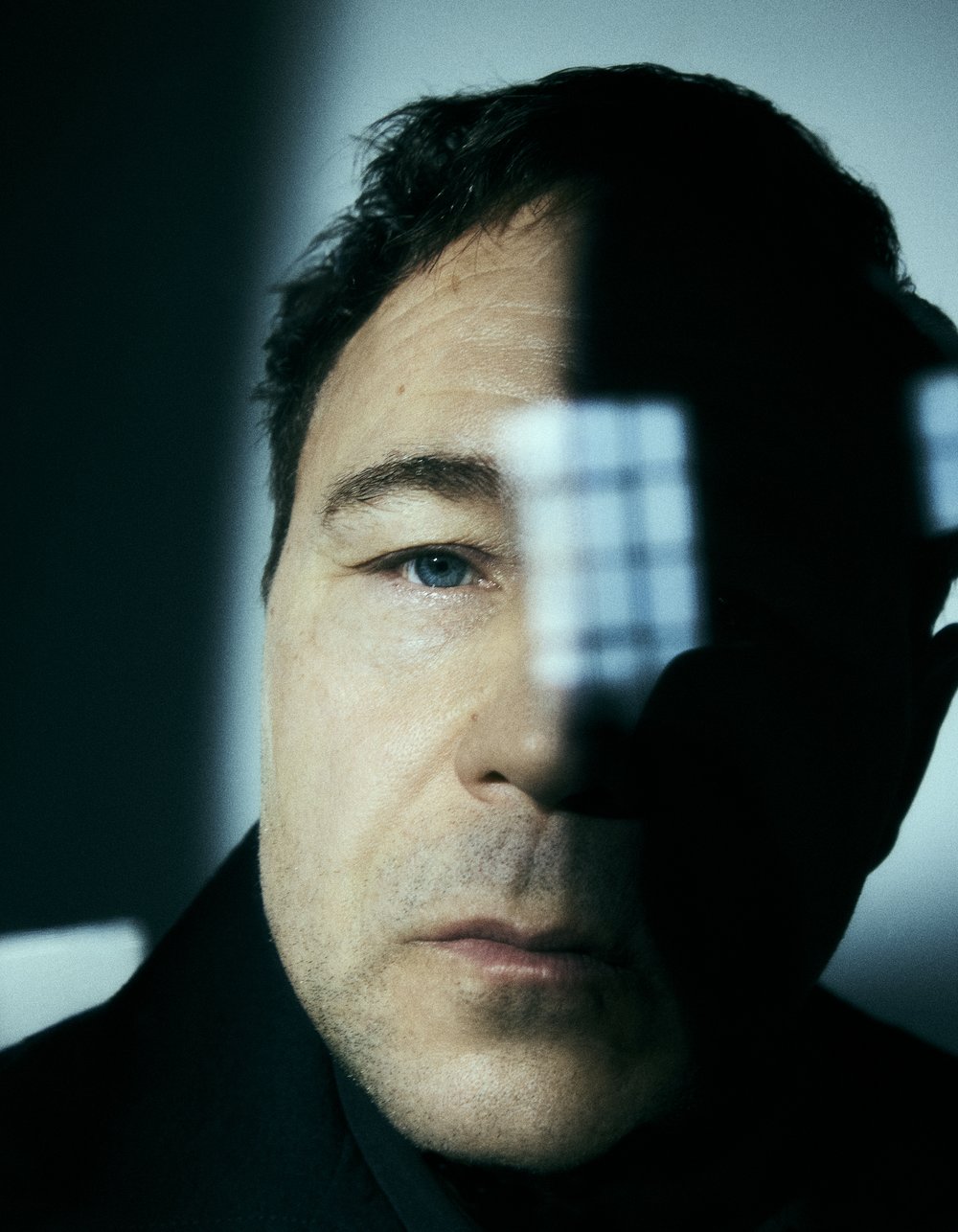 Close-up portrait of a man with dark hair and blue eyes, partially shadowed with a geometric light pattern on a light-blue background.