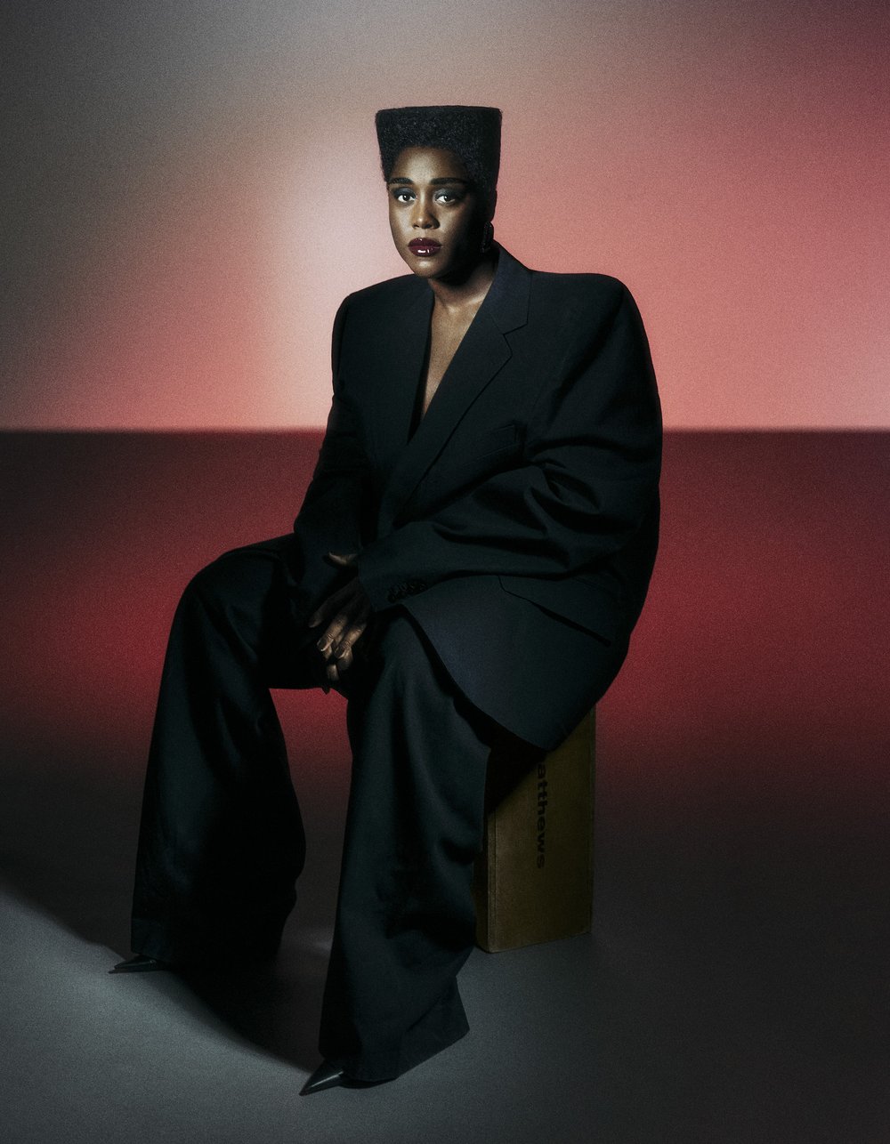 Portrait of a Black woman in a dark suit sitting on a wooden crate in a photo studio with red and white gradient background lighting.