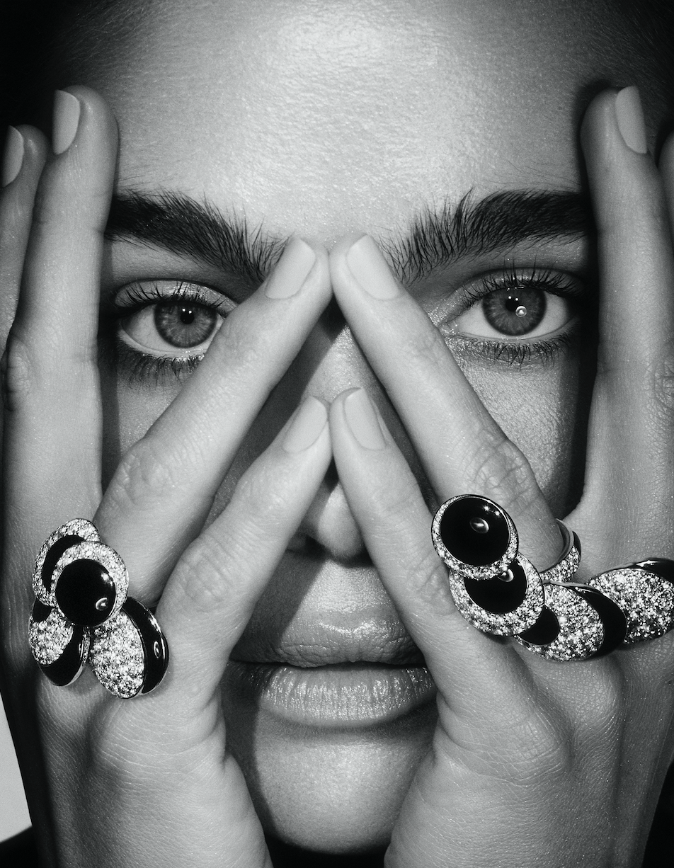 Close-up portrait of a person's face, partially covered by hands wearing rings, emphasizing eyes and jewelry in a studio setting.