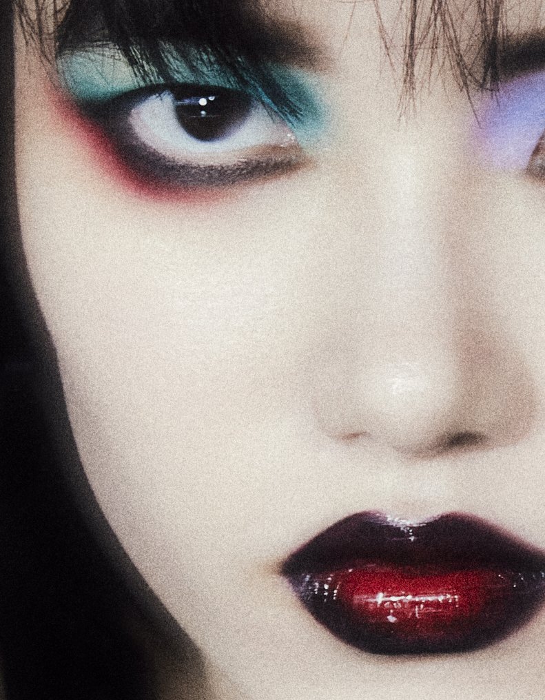 Close-up beauty portrait of a fair-skinned adult woman with dark hair, dramatic eye makeup in teal, red and purple, and dark red glossy lips.