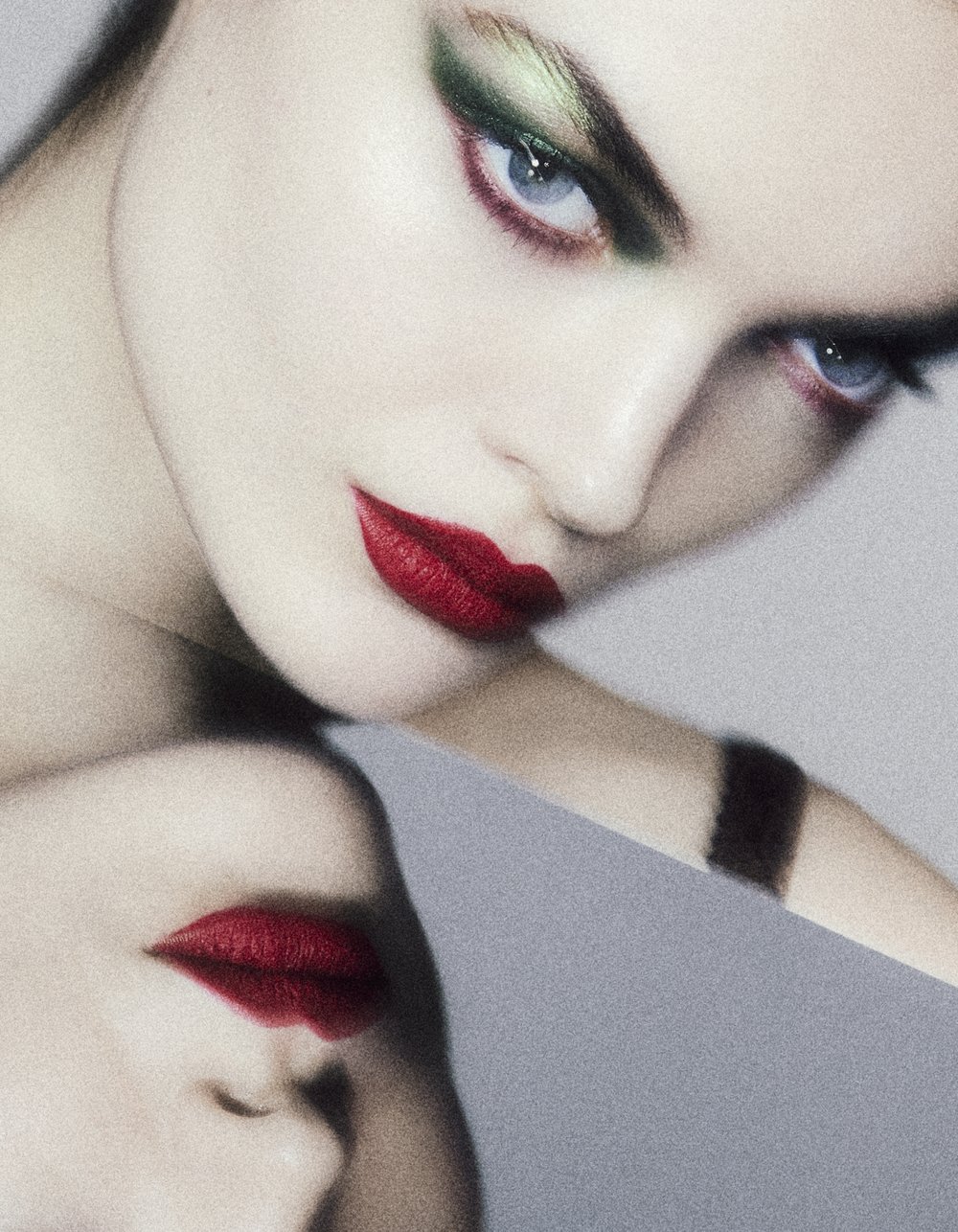 Close-up portrait of a woman with dramatic green and red eye makeup, and striking red lipstick against a smooth grey background.