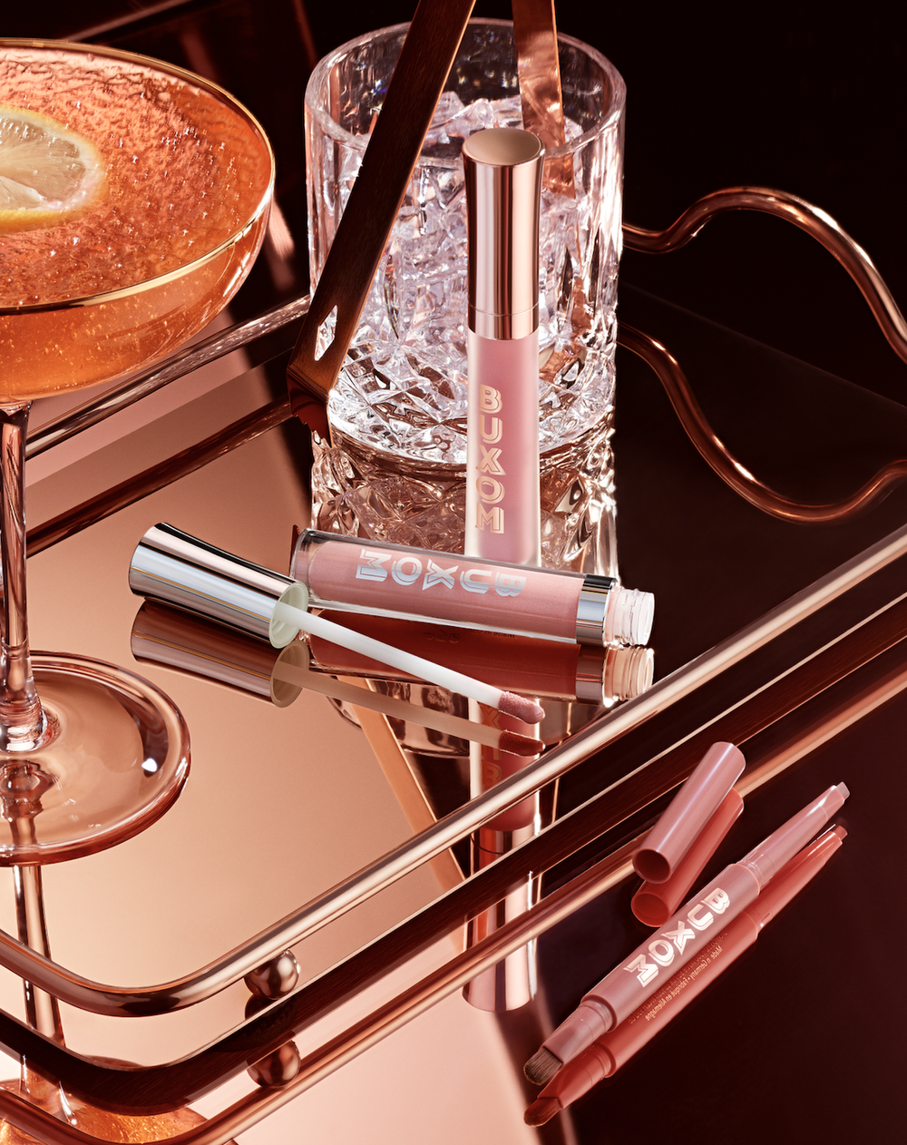Cosmetic still life: Buxom lipsticks, lip liners, and glosses arranged on a reflective gold metal tray with cocktail glass.