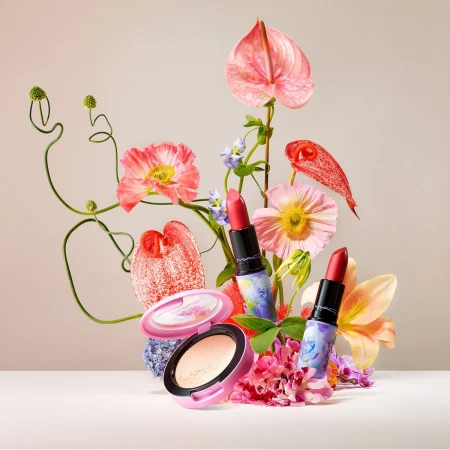 MAC makeup products are arranged among colorful flowers in a bright, soft studio setting for a cosmetic product shot.