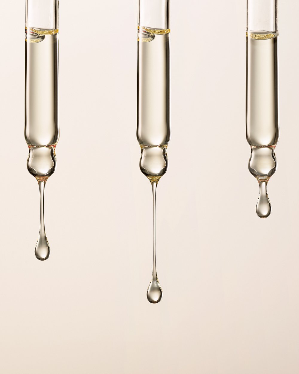 Close-up of three glass pipettes with golden liquid dripping against a light beige backdrop, highlighting the texture and viscosity of the fluid.