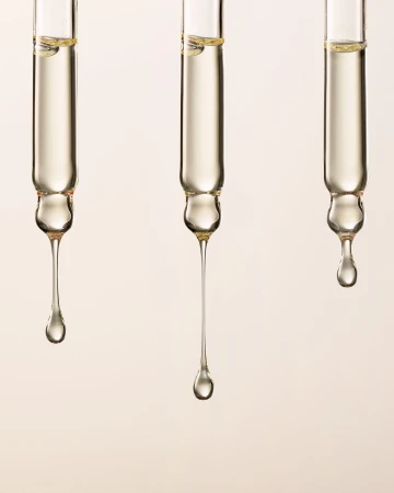 Close-up of three glass pipettes with golden liquid dripping against a light beige backdrop, highlighting the texture and viscosity of the fluid.