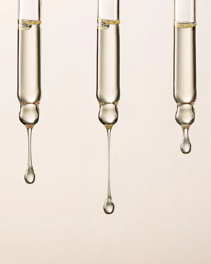 Three glass pipettes with droplets of golden serum suspended against a neutral cream background.