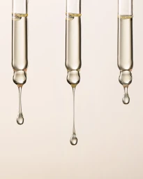 Three glass pipettes with droplets of golden serum suspended against a neutral cream background.