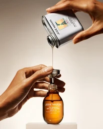Two hands pouring golden argan oil from a silver refill tin through a small metal funnel into a glass bottle.