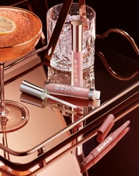 Buxom lip gloss tubes and a lip liner resting on a reflective copper tray beside an iced cocktail and a crystal glass.