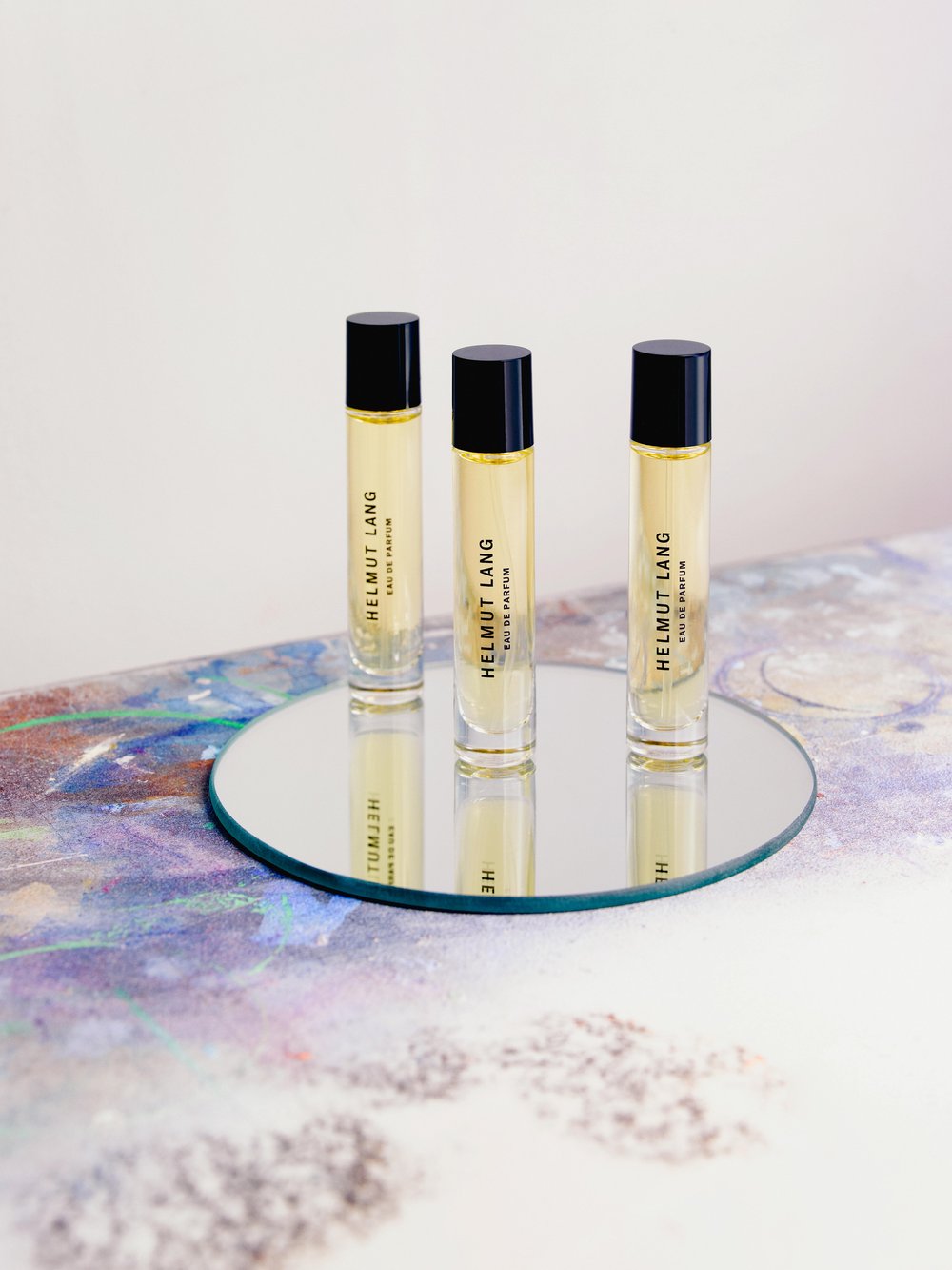 Three Helmut Lang roll-on perfumes displayed on a round mirror against a white background with subtle lavender and blue accents.
