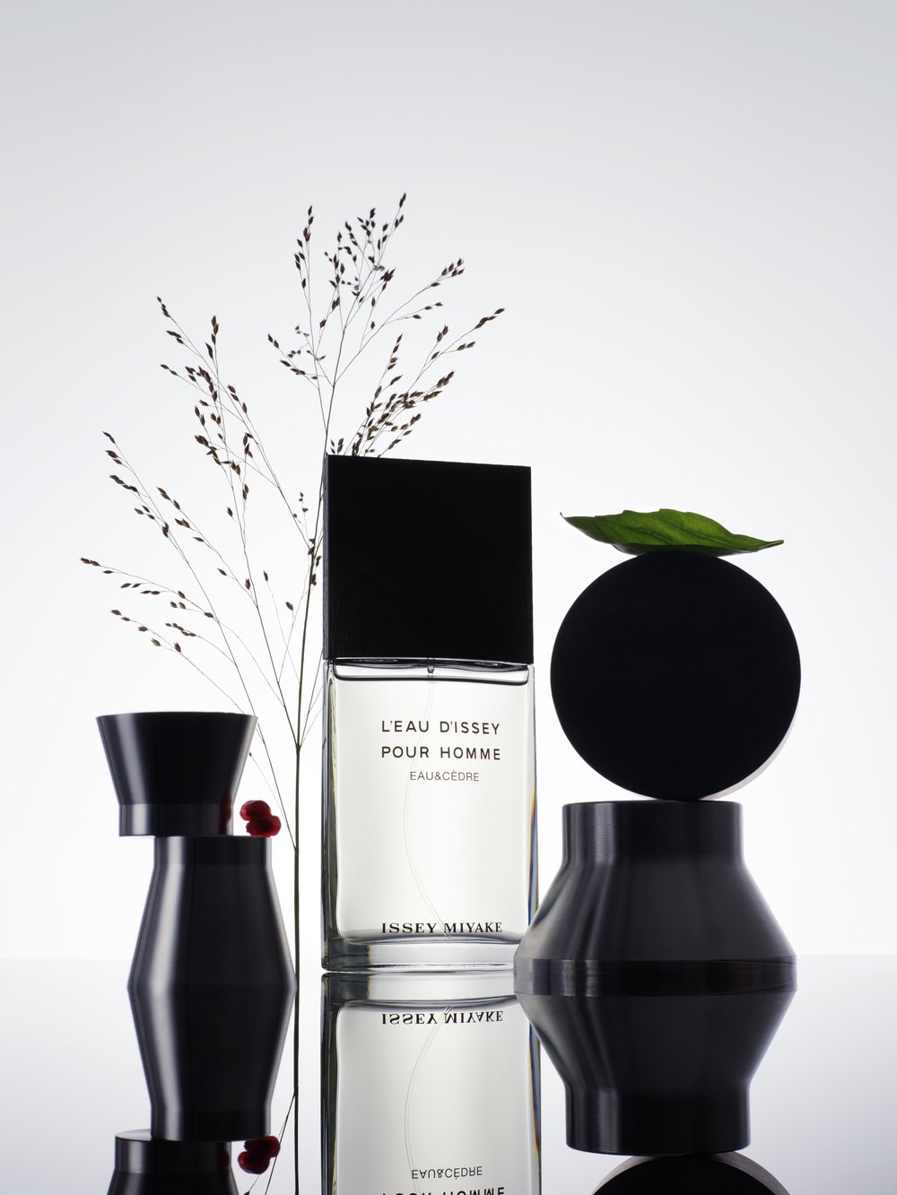Issey Miyake L'Eau d'Issey Pour Homme Eau Cédre fragrance bottle with a black cap, grass, and decorative vases on a reflective surface.