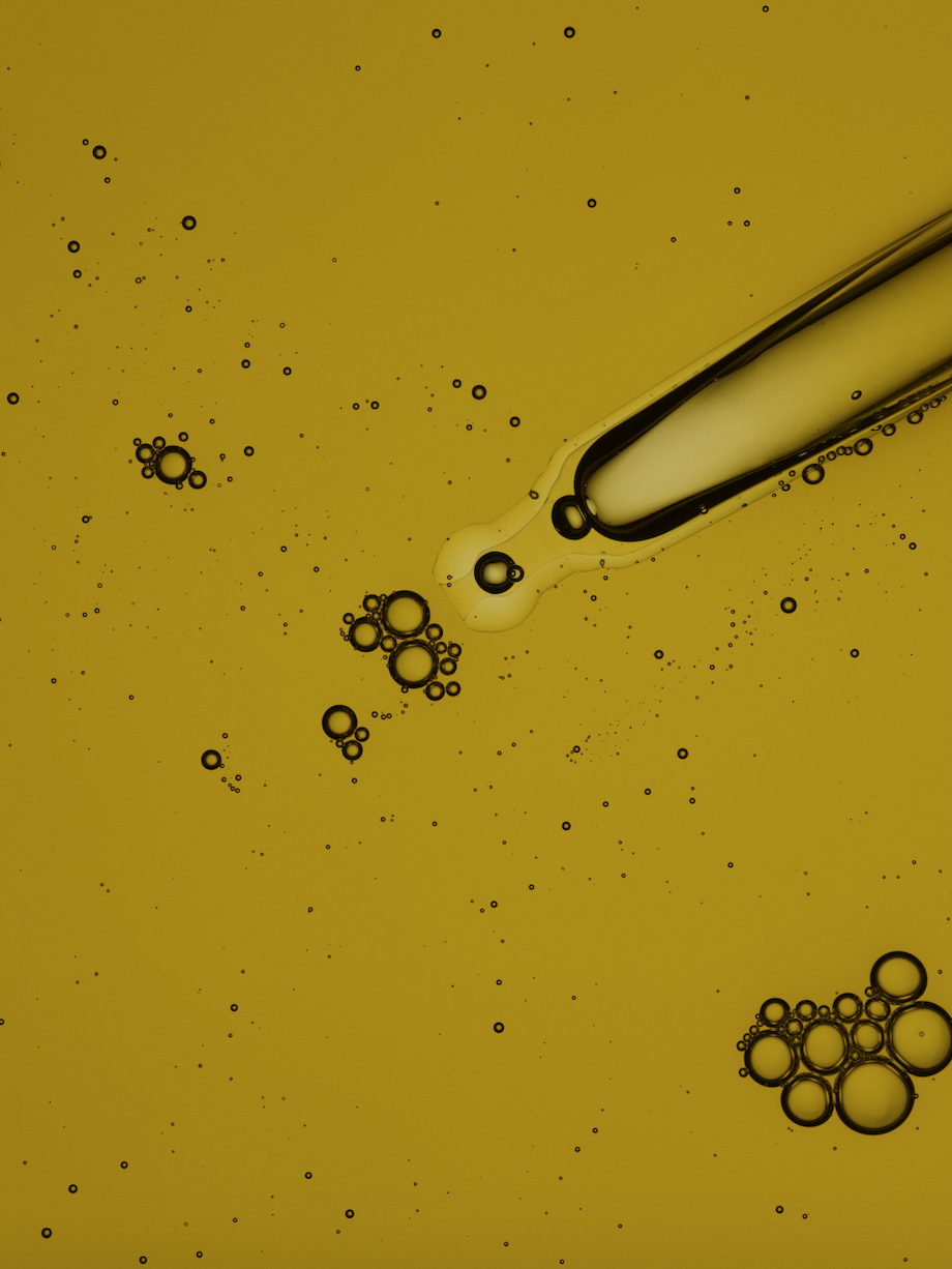 Close-up of a clear glass dropper releasing liquid with bubbles into a yellow liquid solution, set against a seamless backdrop.