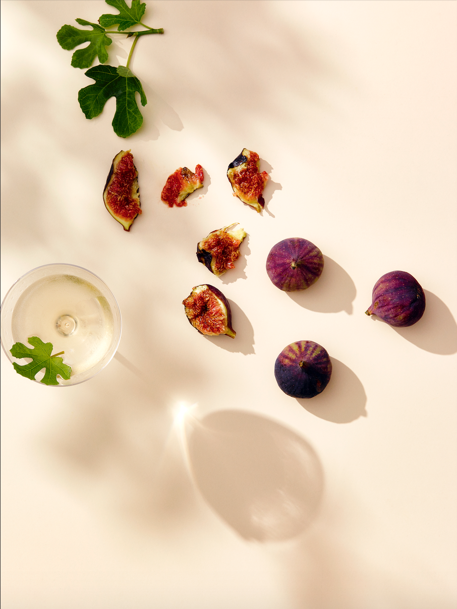 Overhead shot of fresh figs, sliced figs, green fig leaves, and a cocktail in a coupe glass, arranged on a light pink surface.
