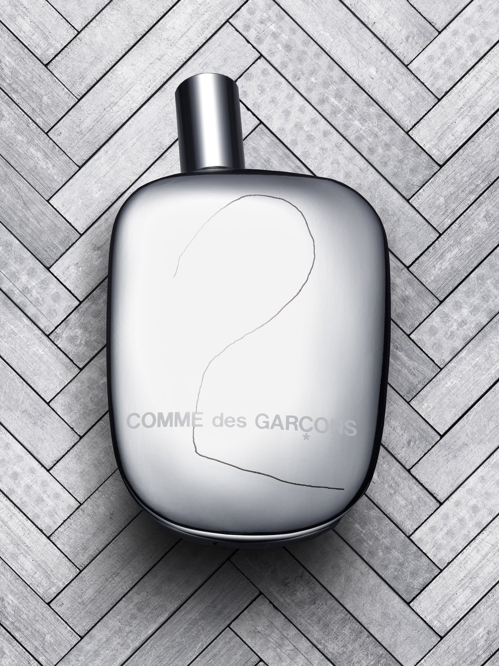 Comme des Garçons perfume bottle on a herringbone wooden surface. Silver container with visible brand name and minimalistic design.