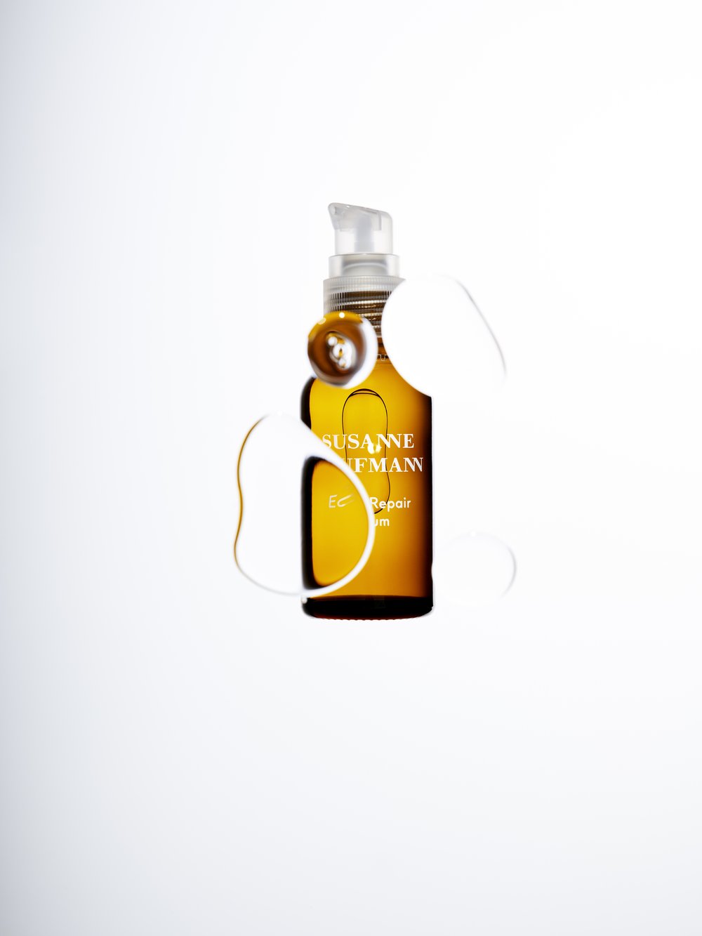Amber glass bottle of Susanne Kaufmann hair repair serum with pump dispenser on a white background with simulated liquid drops.