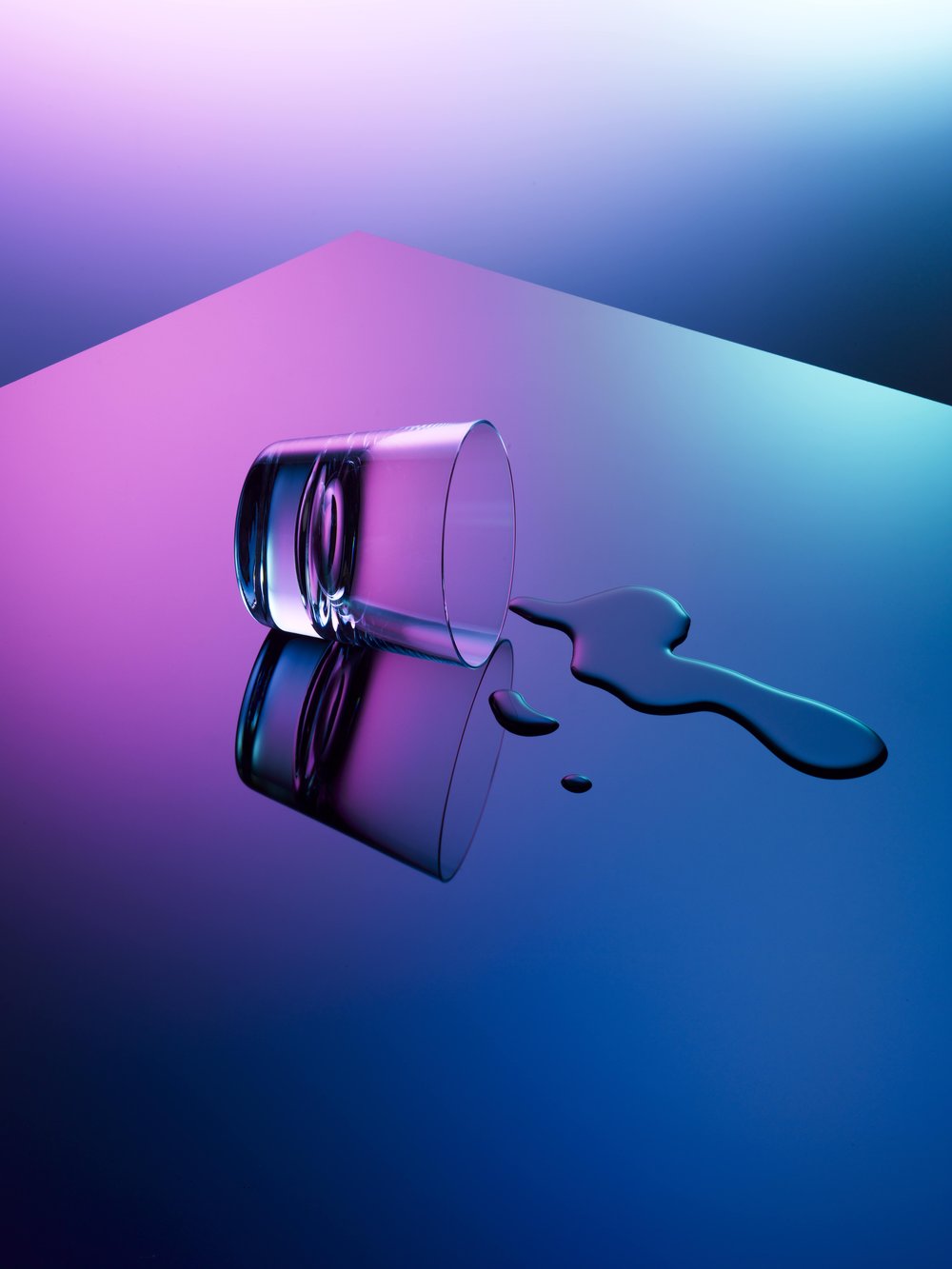 A clear glass laying on its side with a puddle of liquid spills out onto a blue and purple reflective surface.