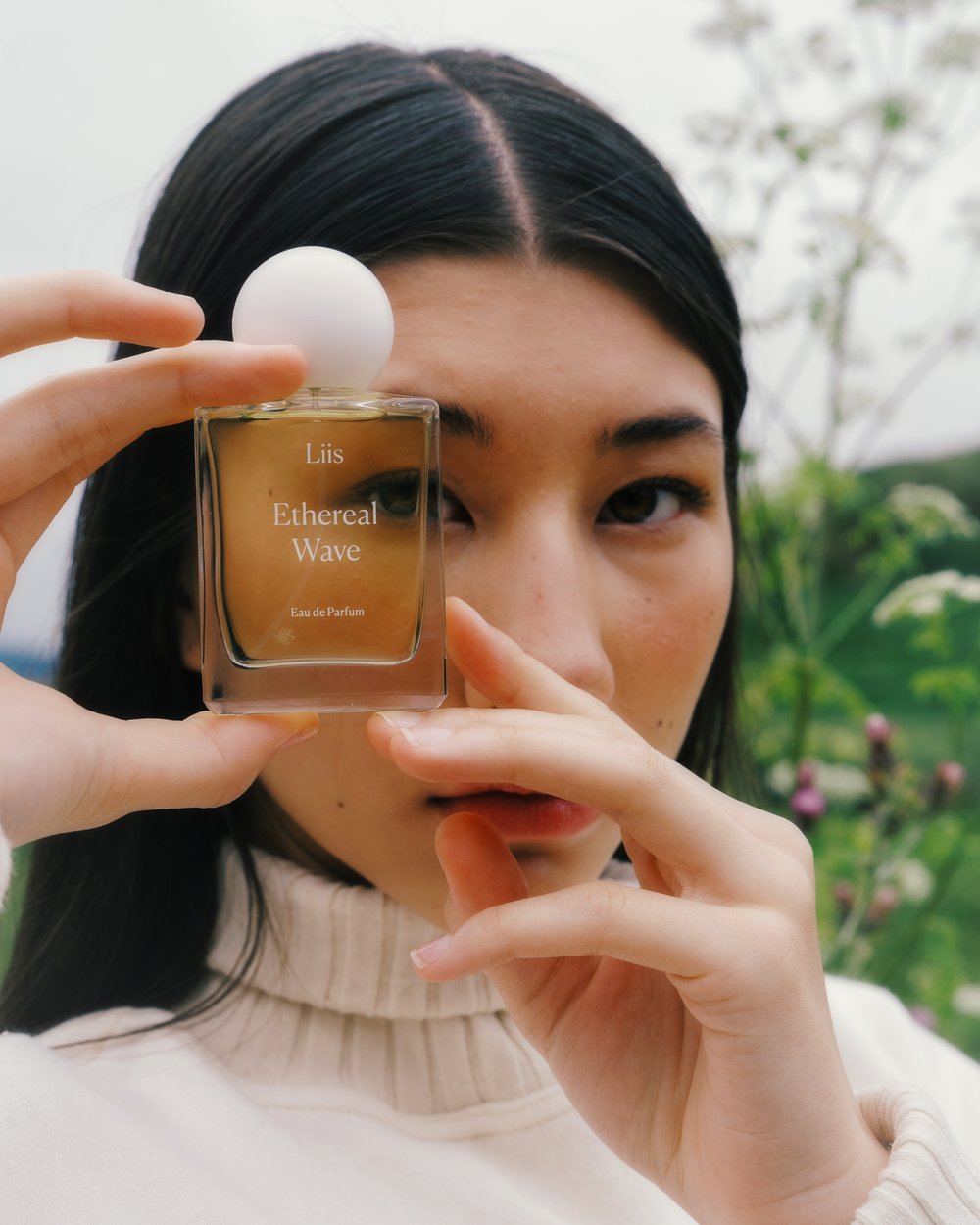 Young woman presents a glass bottle of Ethereal Wave Eau de Parfum by Liis, set against an outdoor background with white flowering plants.