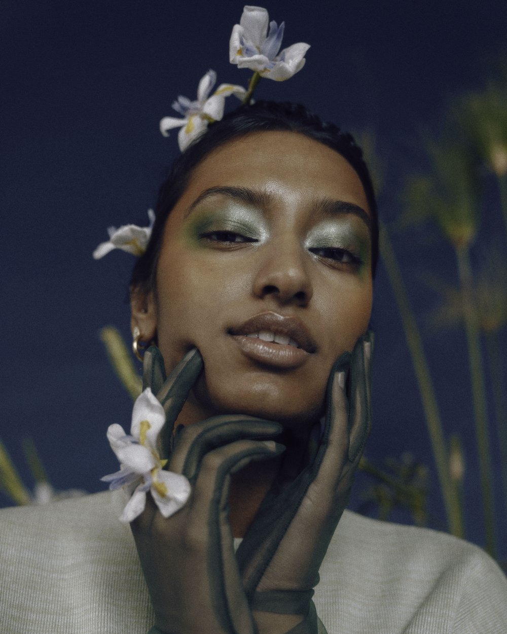 Close-up of a woman with dark hair, olive complexion, and neutral expression wearing sheer green gloves and white flowers in her hair.