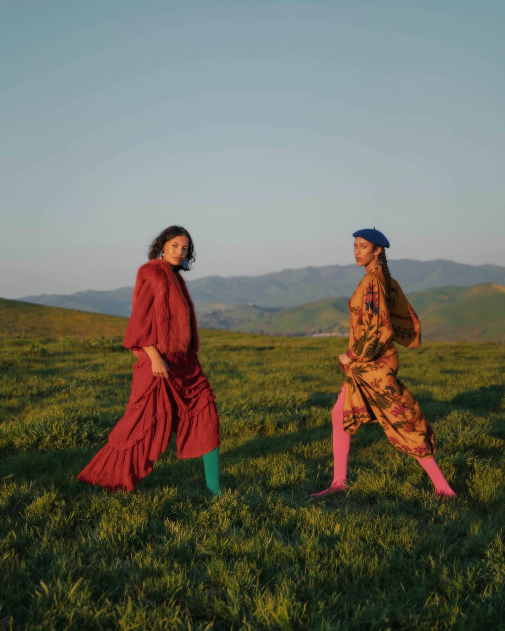 Two women in vintage dresses pose in a grassy field with distant mountains under a blue sky. Fashion editorial outdoor setting.