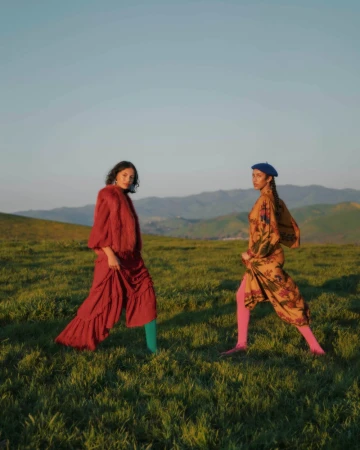 Two women in vintage dresses pose in a grassy field with distant mountains under a blue sky. Fashion editorial outdoor setting.