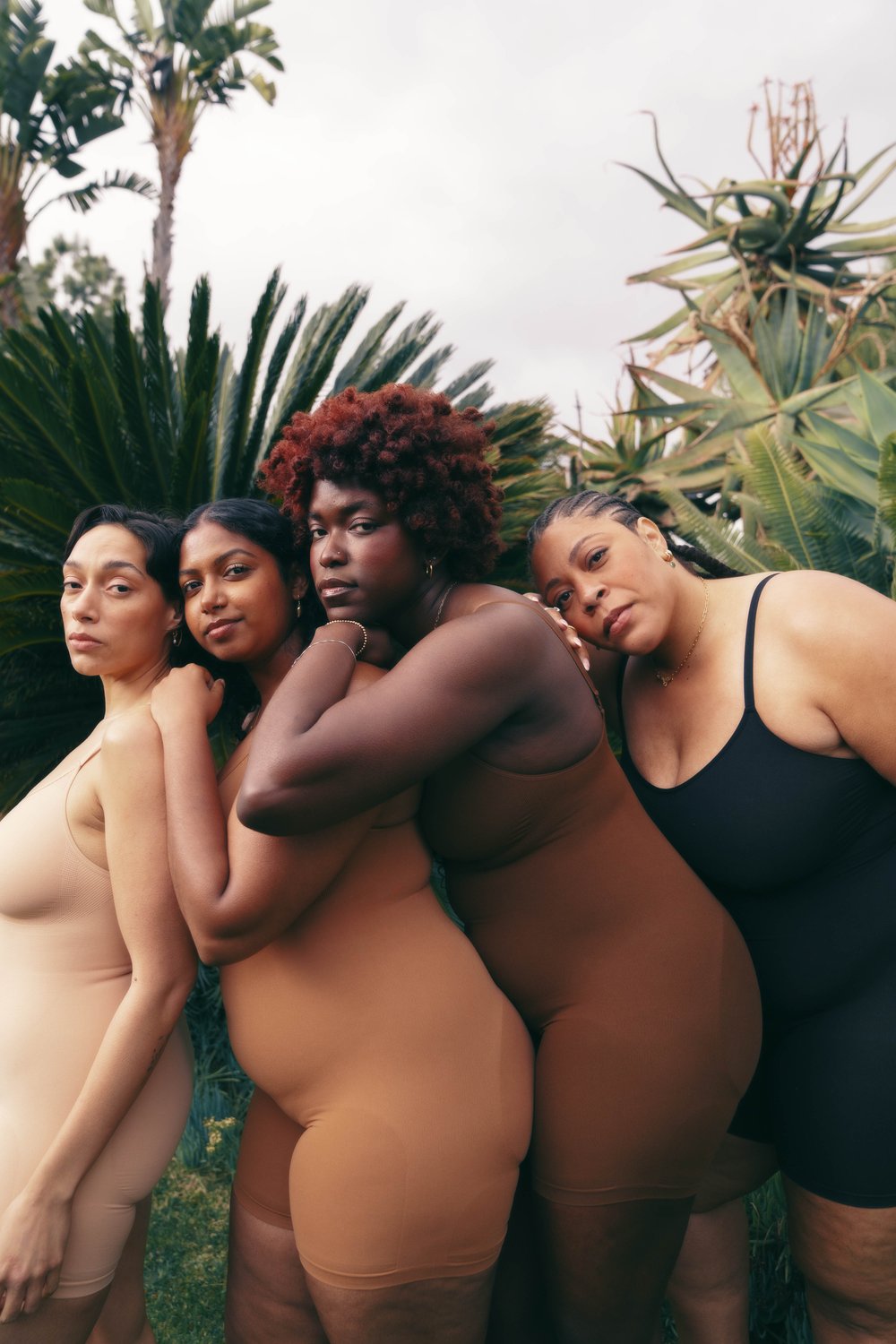 Four women of diverse ethnicities and body types pose together in a garden setting, wearing nude and black bodysuits, showcasing natural beauty.