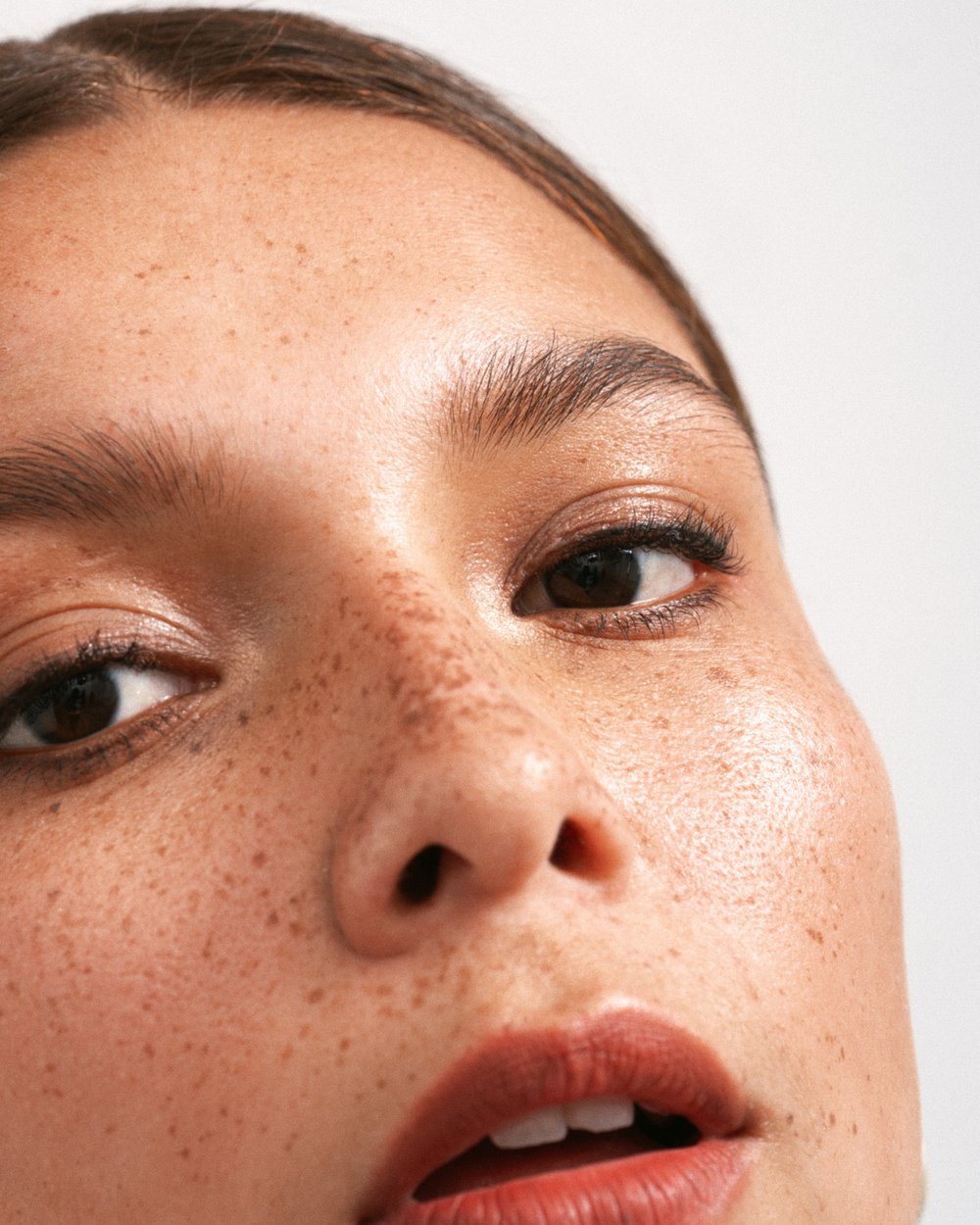 Close-up portrait of a woman's face with freckles, brown eyes, and red lipstick against a clean, bright white background in a beauty context.