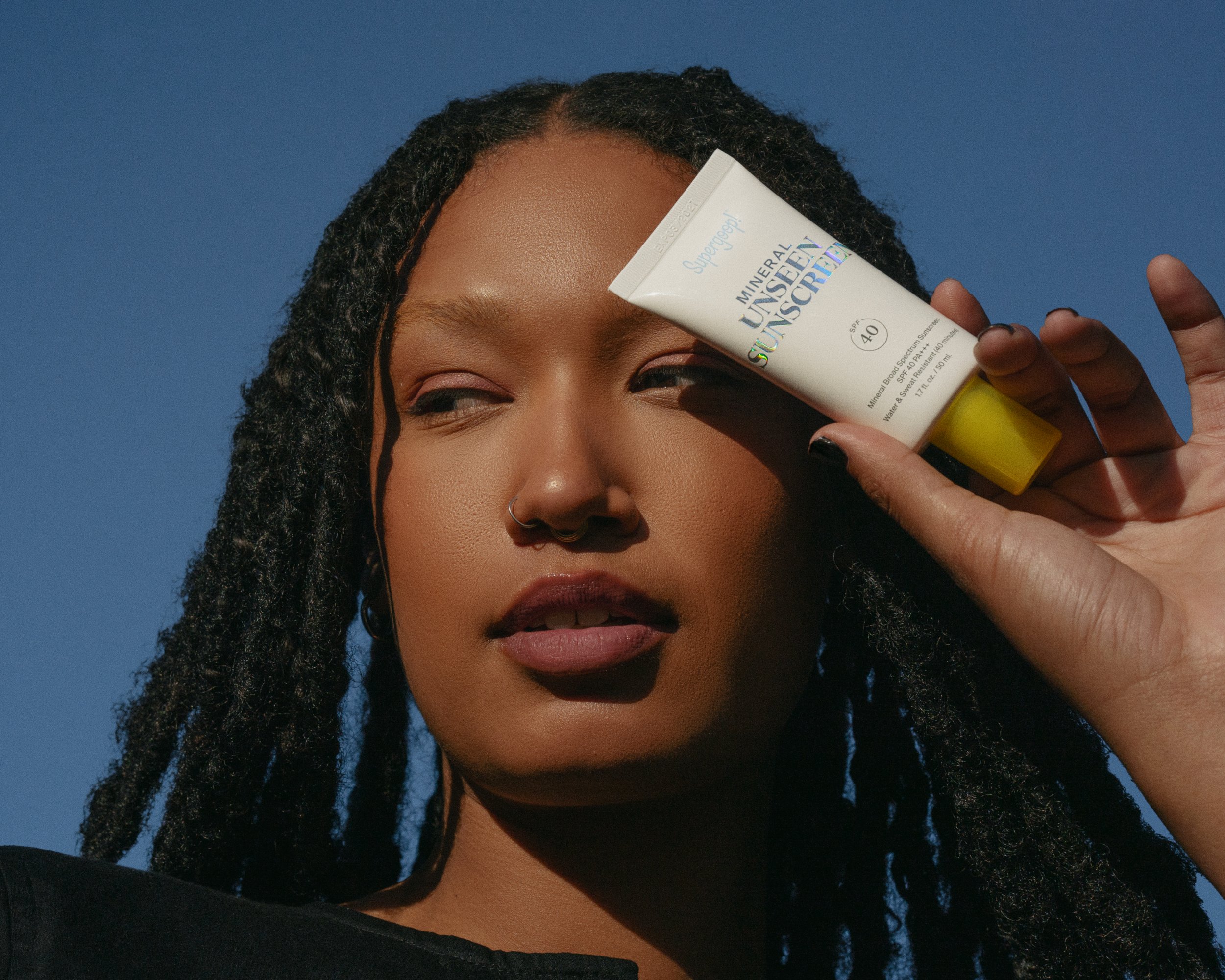 A person with dark skin and dreadlocks holding a tube of white and yellow sunscreen against a blue sky background in an eye-level shot.