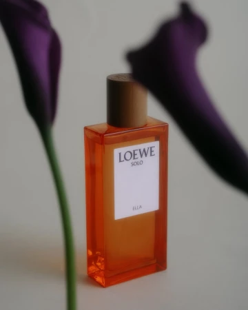 Elegant still life shot of Loewe Solo Ella perfume bottle, brown cap, orange glass, framed by out-of-focus purple calla lilies.