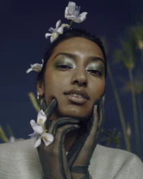 Close-up portrait of a South Asian woman wearing green gloves, shimmering green eyeshadow, and white flowers in her hair by Simrah Farrukh.