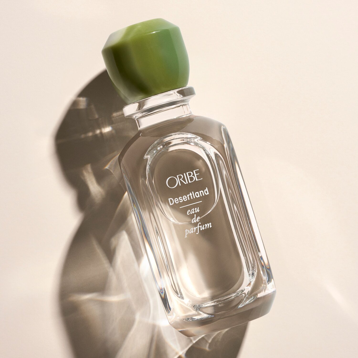 Oribe Desertland eau de parfum: A clear glass bottle with a green cap sits against a soft, neutral backdrop, casting a subtle shadow.