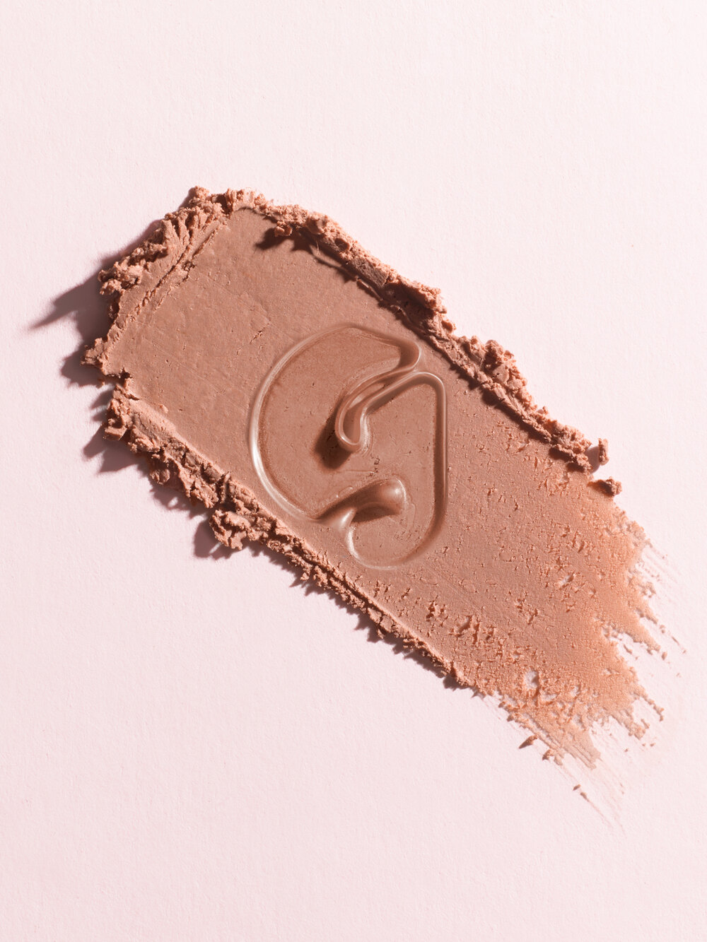 Close-up of a swatch of light brown cosmetic foundation on a light pink background with an embossed 'G' logo.