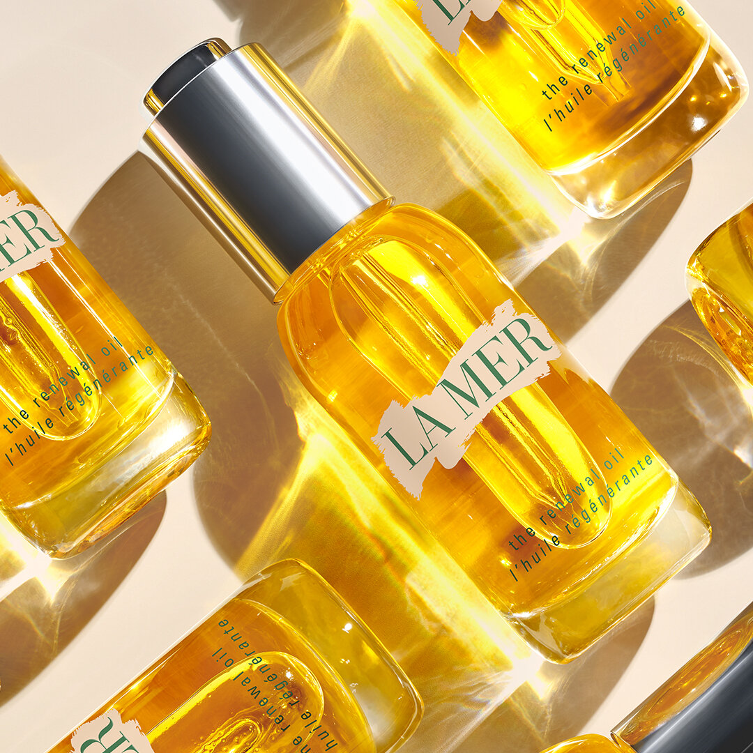 Close-up studio shot of multiple clear glass bottles of La Mer renewal oil with silver lids on a cream colored background.
