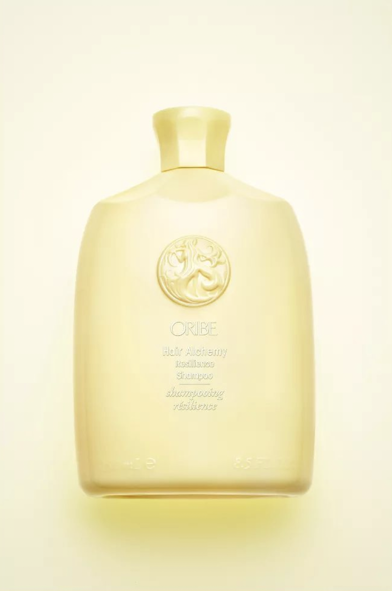 Oribe Gold Lust Nourishing Hair Oil bottle on a light yellow background. The bottle has a gold cap and a circular emblem.