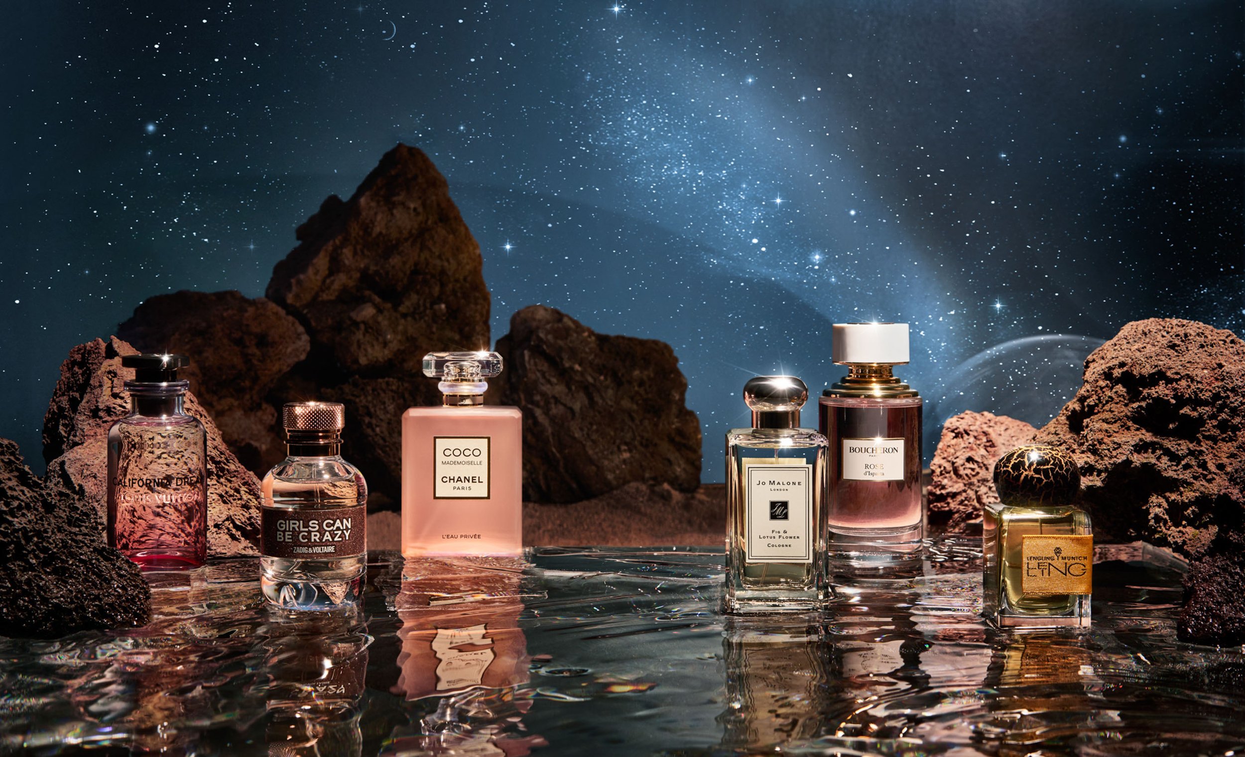 Several perfume bottles are displayed against a space background with dark rocky terrain and water-like reflective surface in a studio setting.
