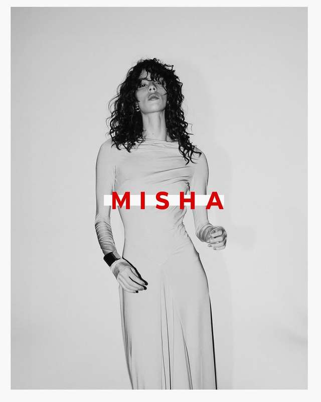 A slender woman with long curly hair in a long-sleeved dress poses in a studio setting, with the word MISHA overlaid on the frame.
