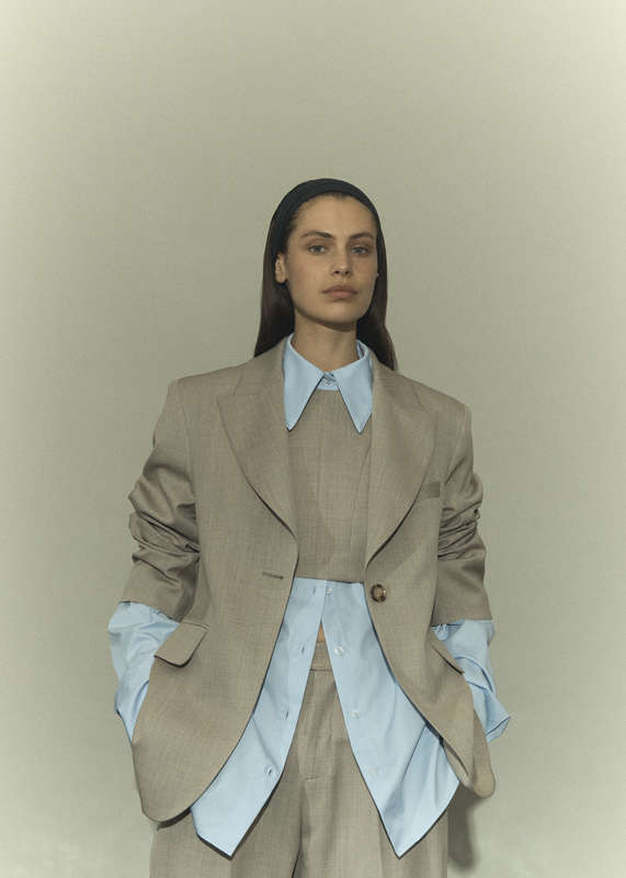 Fashion portrait of a woman in a light brown suit with a blue collared shirt, headband, and light skin, posing indoors.